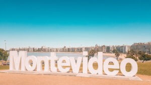 The image shows large, white letters spelling out "Montevideo" against the backdrop of a coastal cityscape. The bright blue sky and the waterfront, with modern buildings lining the shore, highlight the beauty of Montevideo, Uruguay's capital.