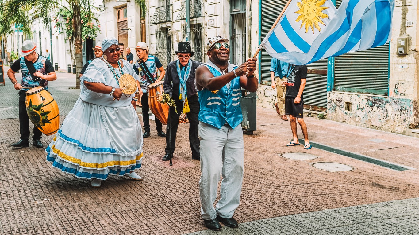 The image captures a vibrant street parade in Montevideo, Uruguay. Participants are dressed in colorful, traditional attire, with one man waving a large Uruguayan flag. Musicians play drums and other instruments, creating a lively and festive atmosphere in the urban setting.