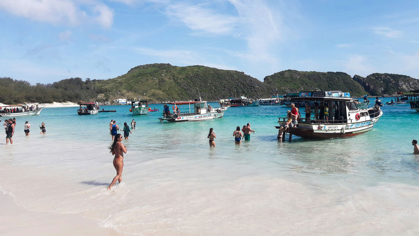 Tourists enjoy the clear turquoise waters at Farol Beach in Arraial do Cabo, Brazil. Multiple boats are moored near the shore, with people disembarking into the shallow water. The scene captures a bustling day at one of the most popular beaches in Arraial do Cabo, surrounded by lush green hills under a bright blue sky.