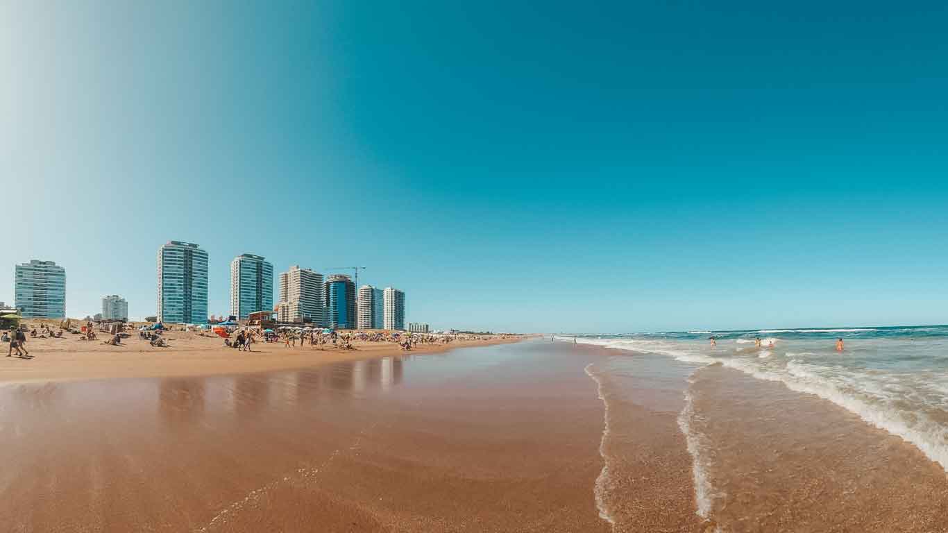 The image shows Playa Brava in Punta del Este, Uruguay, with its expansive sandy beach and modern high-rise buildings lining the shore. Beach goers can be seen enjoying the sunny weather, with some people in the water and others relaxing on the sand, set against a clear blue sky.