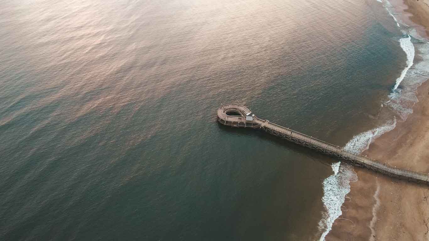 Aerial view of Playa Mansa, showing a curved pier stretching into the calm ocean waters. The coast features gentle waves lapping the beach, creating a serene coastal setting that can be discovered on a stroll, making it one of the best things to do in Punta del Este.