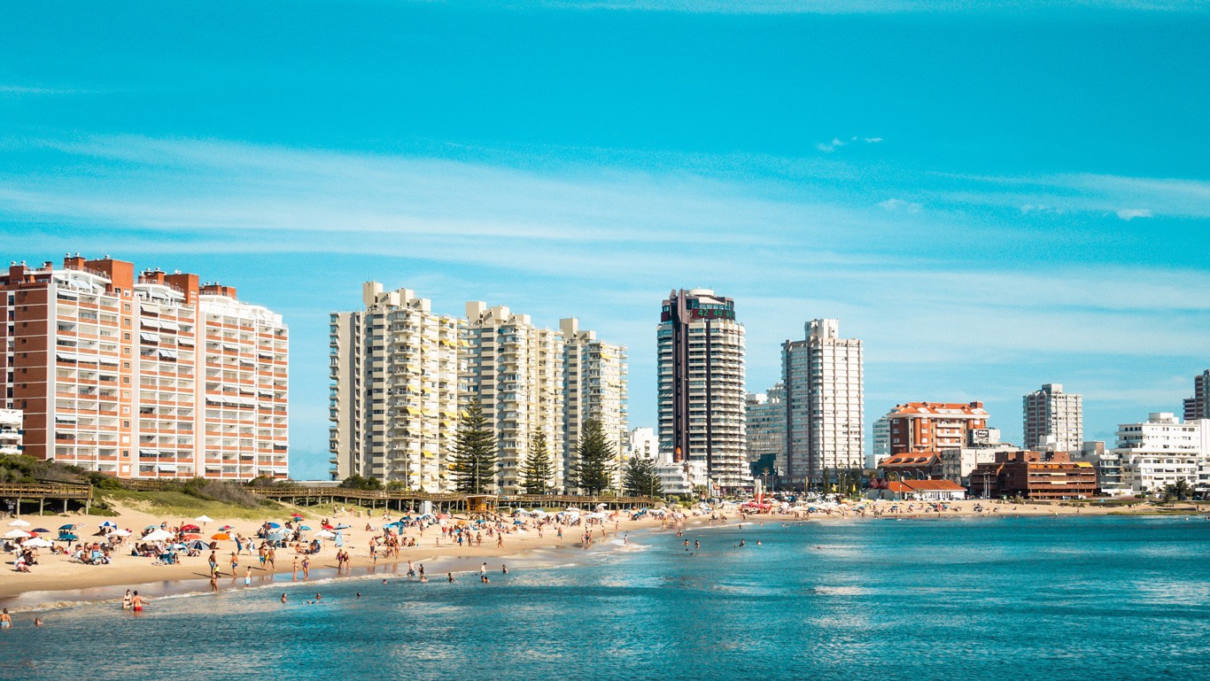 This image shows a vibrant beach scene in Punta del Este, Uruguay. The shoreline is dotted with people enjoying the sun and sea, with colorful umbrellas providing shade. In the background, high-rise buildings tower above the beach, creating a striking contrast against the clear blue sky.