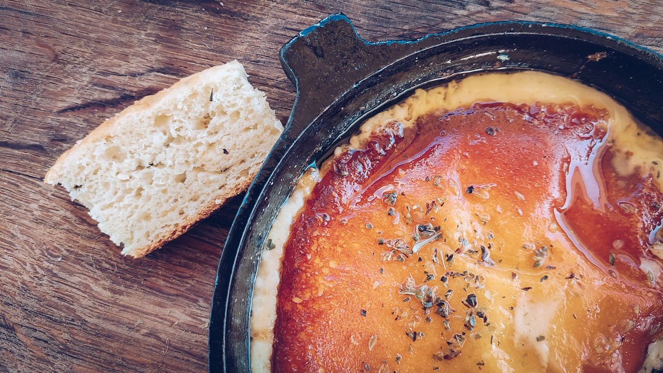 A close-up of a traditional Argentine dish, Provolleta, served in a cast-iron skillet. The melted cheese is topped with herbs and spices, accompanied by a slice of rustic bread, all set on a wooden table.