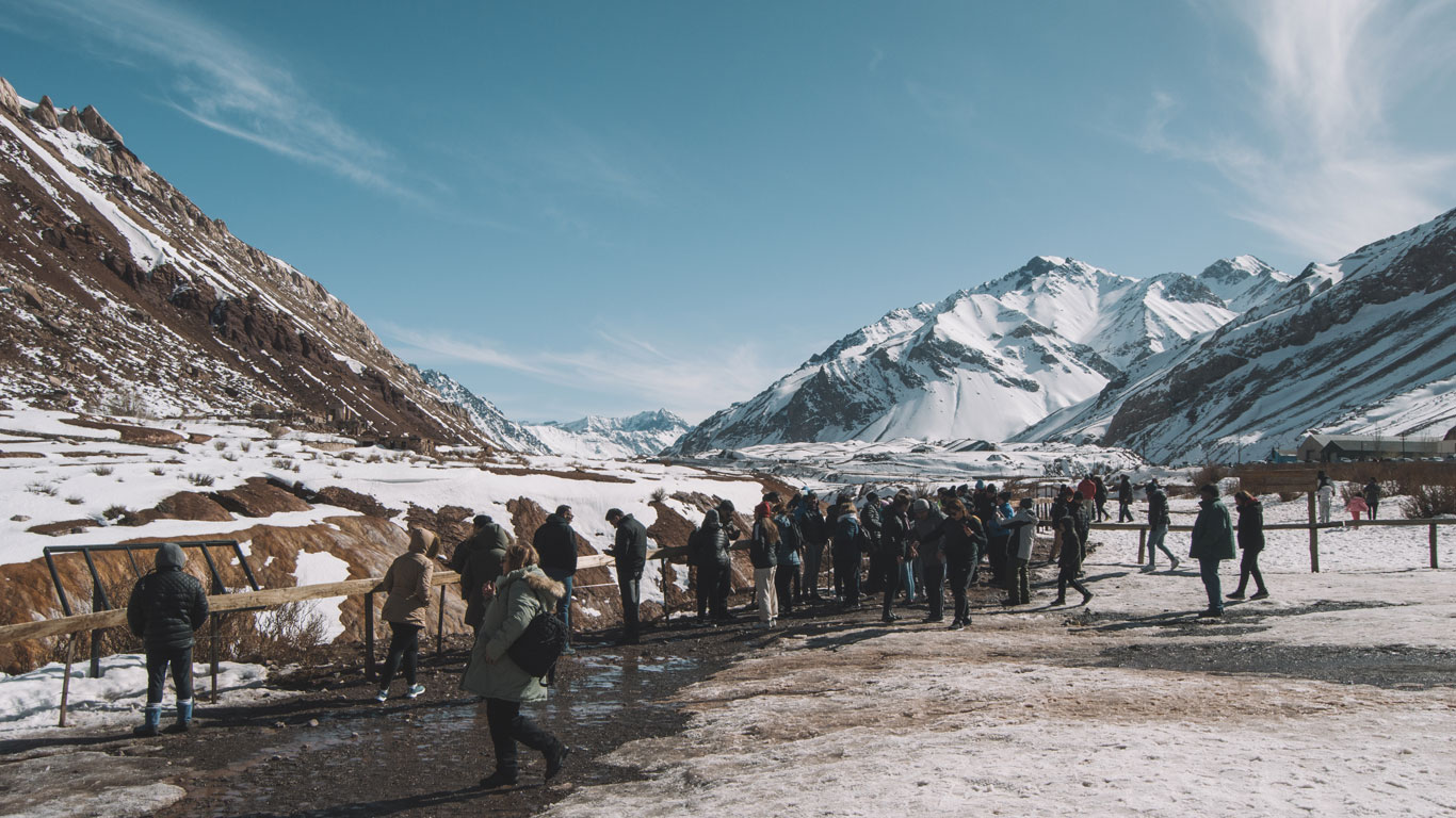 A group of people bundled in winter coats walk along a snowy path at Puente del Inca in Mendoza. The landscape features rugged, snow-covered mountains under a clear blue sky, with the visitors exploring the scenic and historic site.