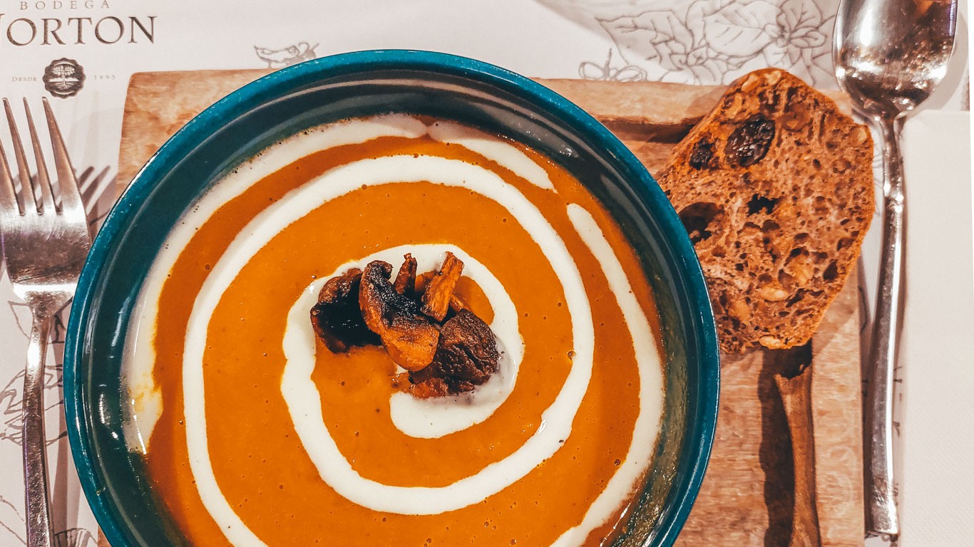 Close-up of a vibrant orange carrot soup served at La Vid Restaurant, Bodega Norton, Mendoza. The soup is elegantly garnished with a swirl of cream and roasted mushrooms and pumpkin seeds in the center, accompanied by a slice of multi-grain bread on a wooden board. The table setting includes silverware and a napkin, all under a soft dining light.