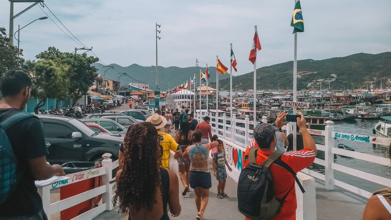 The image captures a lively scene at the Marina dos Pescadores in Arraial do Cabo, filled with tourists and locals. People are walking along a white pier adorned with multiple national flags, heading towards the bustling marina where numerous boats are docked. The area is vibrant and busy, set against a backdrop of green hills and a clear sky, conveying the popular and festive atmosphere of this tourist destination.