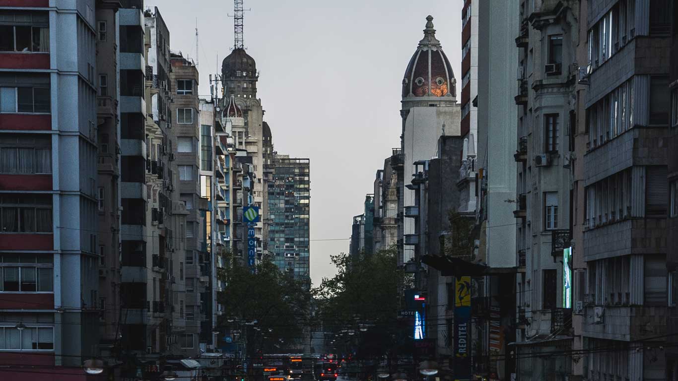 A view of Avenida 18 de Julio in Montevideo, featuring a mix of modern and historic buildings lining the street. The skyline is highlighted by distinctive domed towers, with bustling street activity below, captured during the early evening.