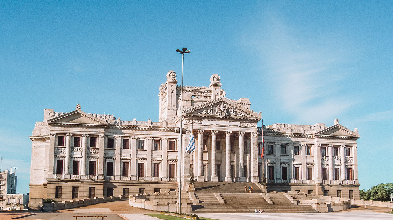 The grand facade of the Palacio Legislativo in Montevideo, featuring neoclassical architecture with towering columns and intricate stone carvings. The Uruguayan flag flies prominently in front of the building, and a few people are seen walking up the steps and around the plaza.