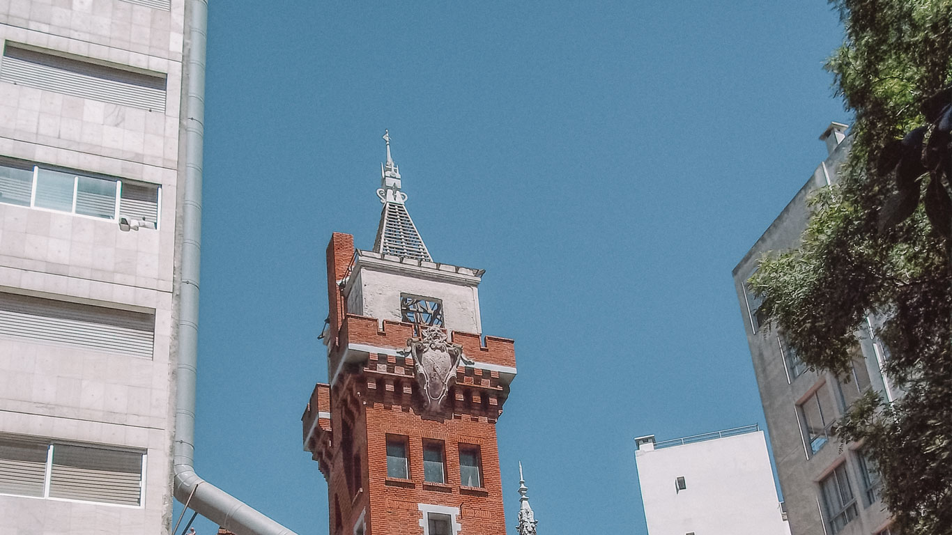 The upper section of Castillo Pittamiglio in Montevideo, featuring its distinct red brick and white stone architecture. The tower is adorned with ornate decorations and topped with a unique spire, contrasting with modern buildings on either side. The bright blue sky provides a vivid backdrop to this historic structure.