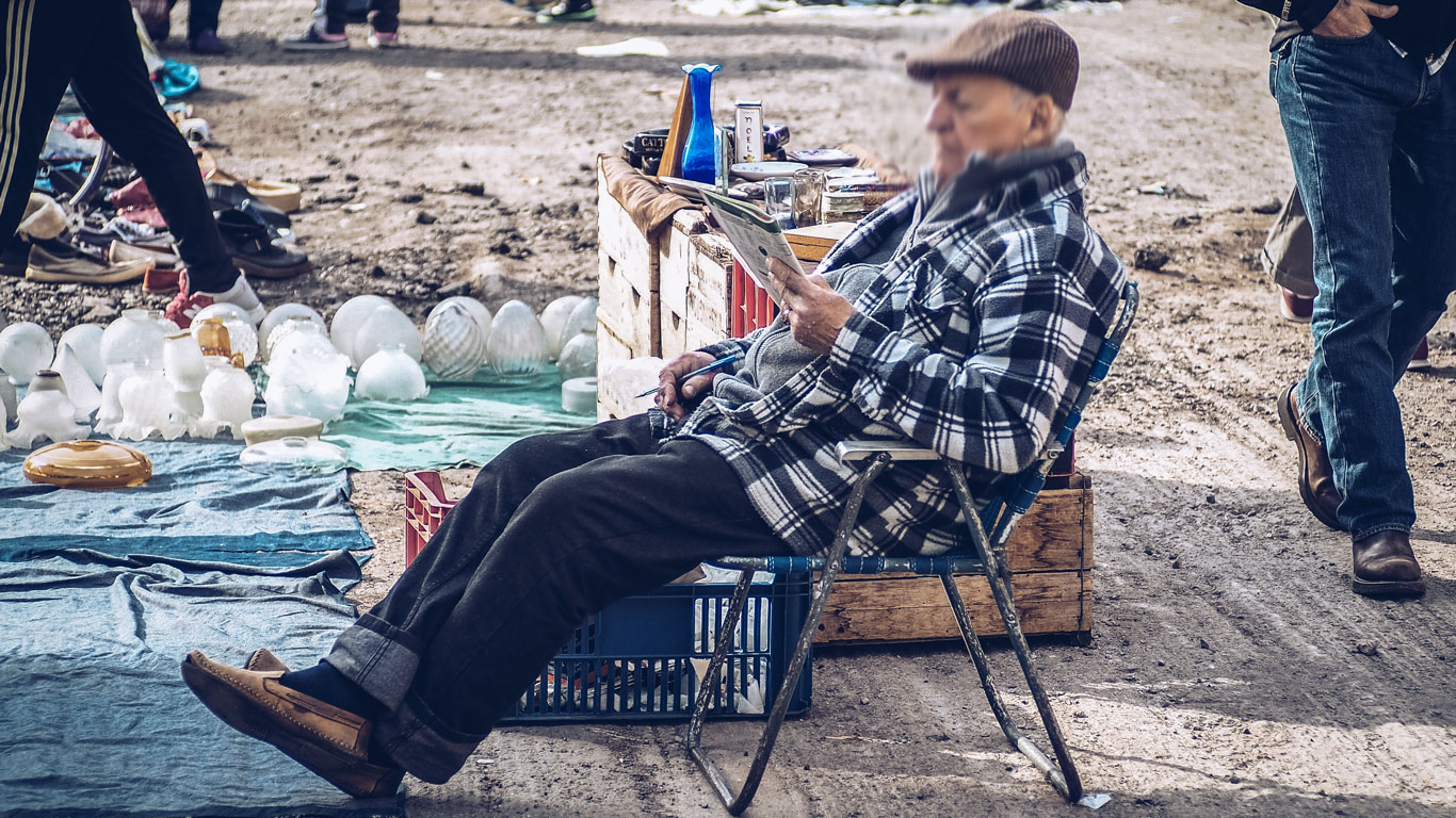 An elderly man in a plaid jacket and flat cap sits in a folding chair, reading a newspaper at the Feria Tristán Narvaja in Montevideo. He is surrounded by various items for sale, including glass lampshades and other trinkets laid out on blankets and wooden crates on the ground. 