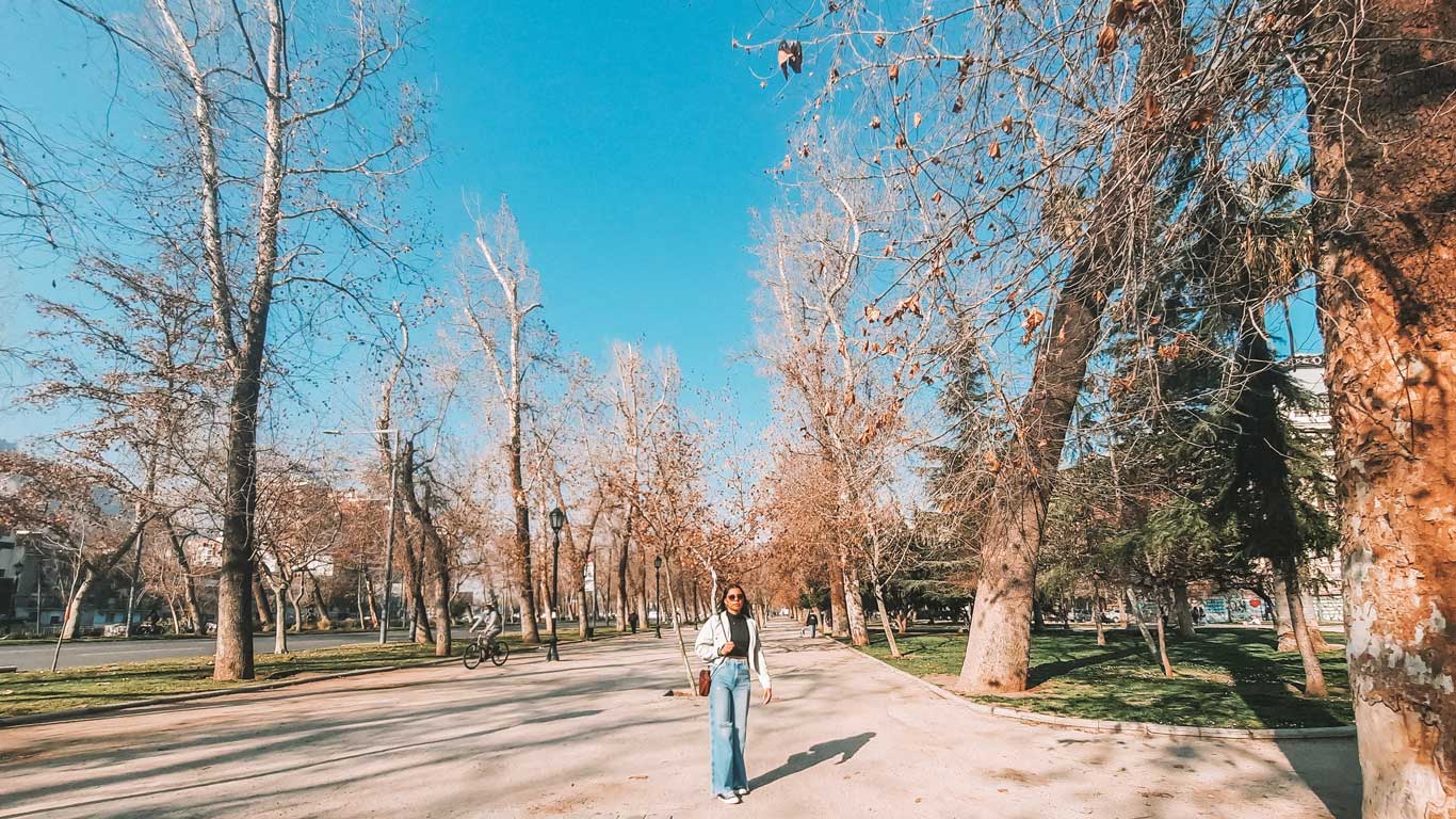 A peaceful park scene in the Bellas Artes neighborhood of Santiago, Chile, featuring a woman walking along a tree-lined path on a sunny day. The bare trees suggest a winter or early spring setting, and the clear blue sky adds a serene backdrop to the tranquil environment.