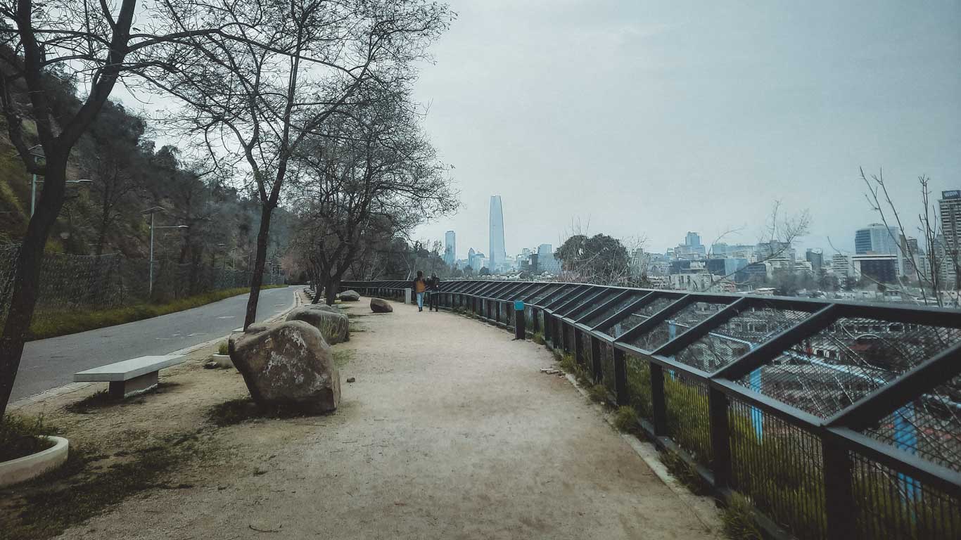 A serene pathway in the Bellavista neighborhood of Santiago, Chile, featuring a pedestrian path lined with trees and benches, with a view of the city's skyline in the distance. The overcast sky and bare trees suggest a chilly day, with the iconic Gran Torre Santiago skyscraper visible in the background.