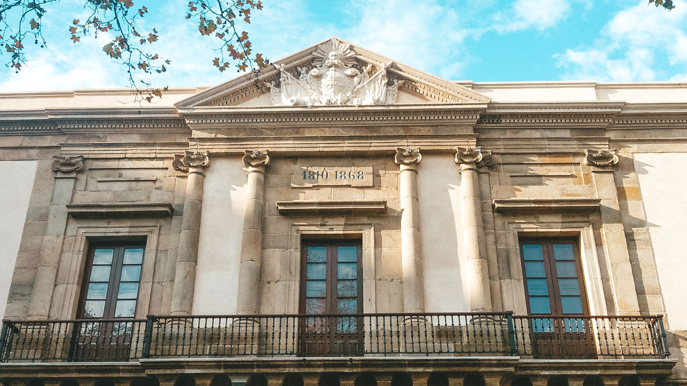 The facade of the Cabildo of Montevideo, featuring neoclassical architectural elements including tall columns and decorative stonework. The building is marked with the years "1810" and "1868" above the entrance, under a clear blue sky with leaves framing the view.