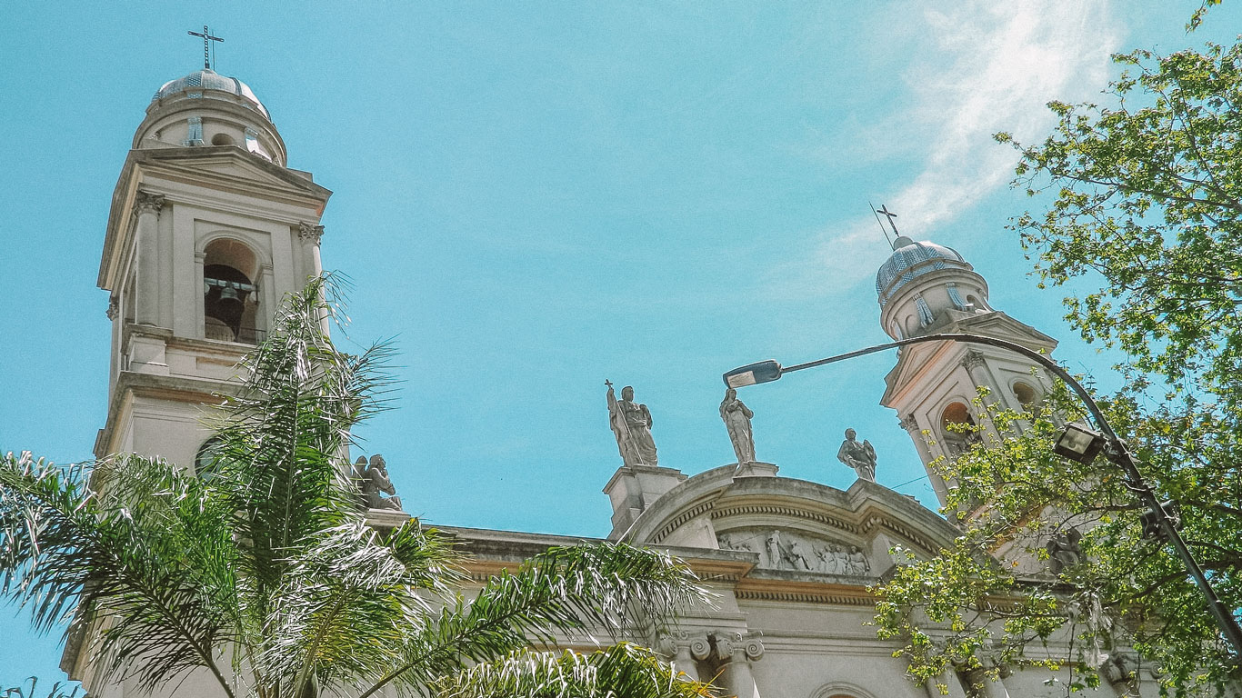 The front view of Catedral Metropolitana in Montevideo, showcasing its twin bell towers and ornate statues atop the facade. The scene is bright and clear, with palm trees and greenery adding to the vibrant atmosphere under a sunny sky.