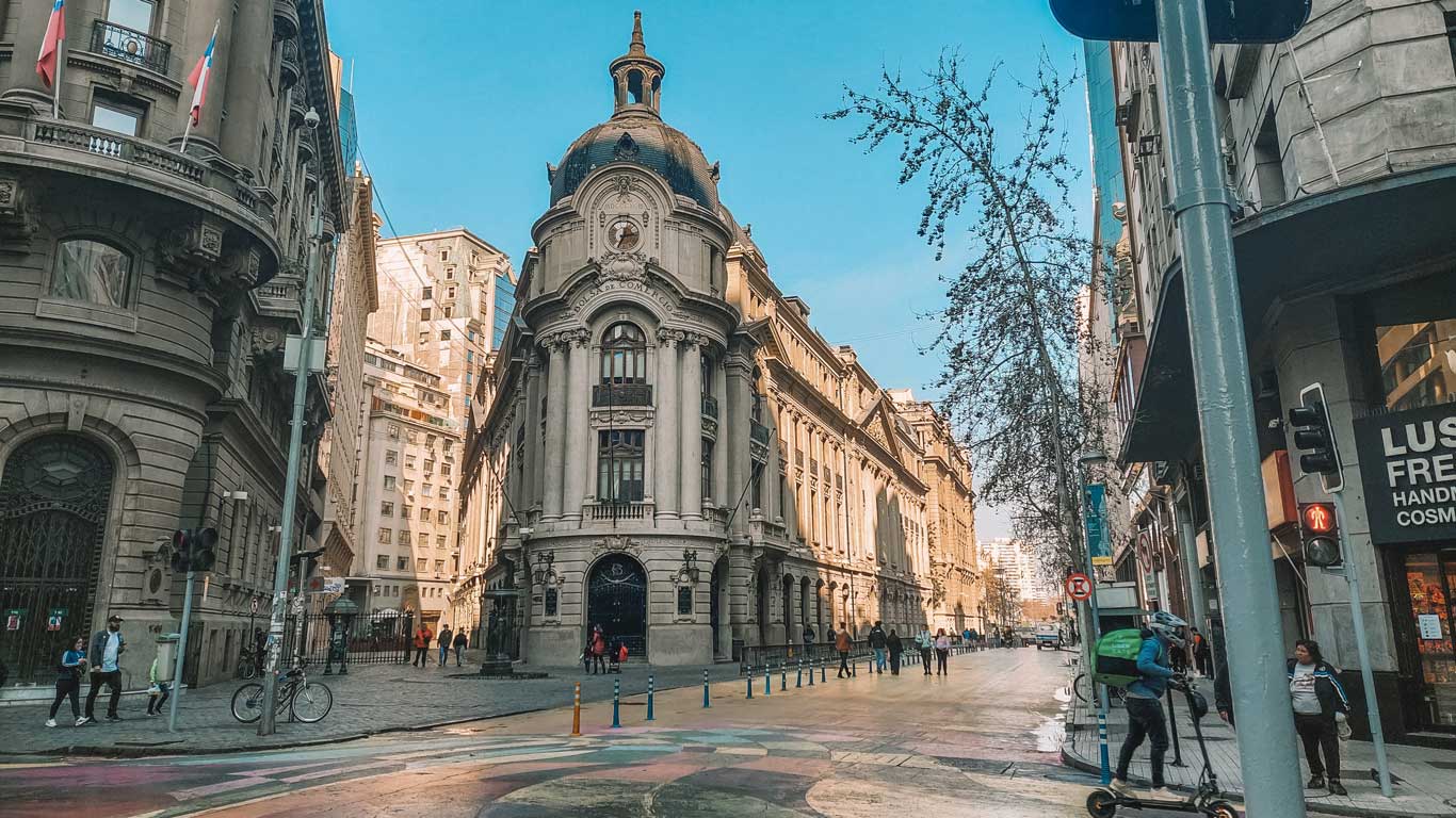 A bustling street in downtown Santiago, Chile, featuring historic architecture with a prominent building marked "Bolsa de Comercio" at the center. The street is lively with pedestrians, cyclists, and a delivery person on a scooter. The scene is accented by the clear blue sky and a mix of modern and classic buildings.