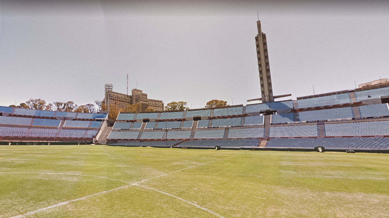 A view of Estadio Centenario in Montevideo, Uruguay, showcasing its expansive seating areas and central field under a clear sky. The iconic tower stands prominently in the background, flanked by surrounding buildings and trees.