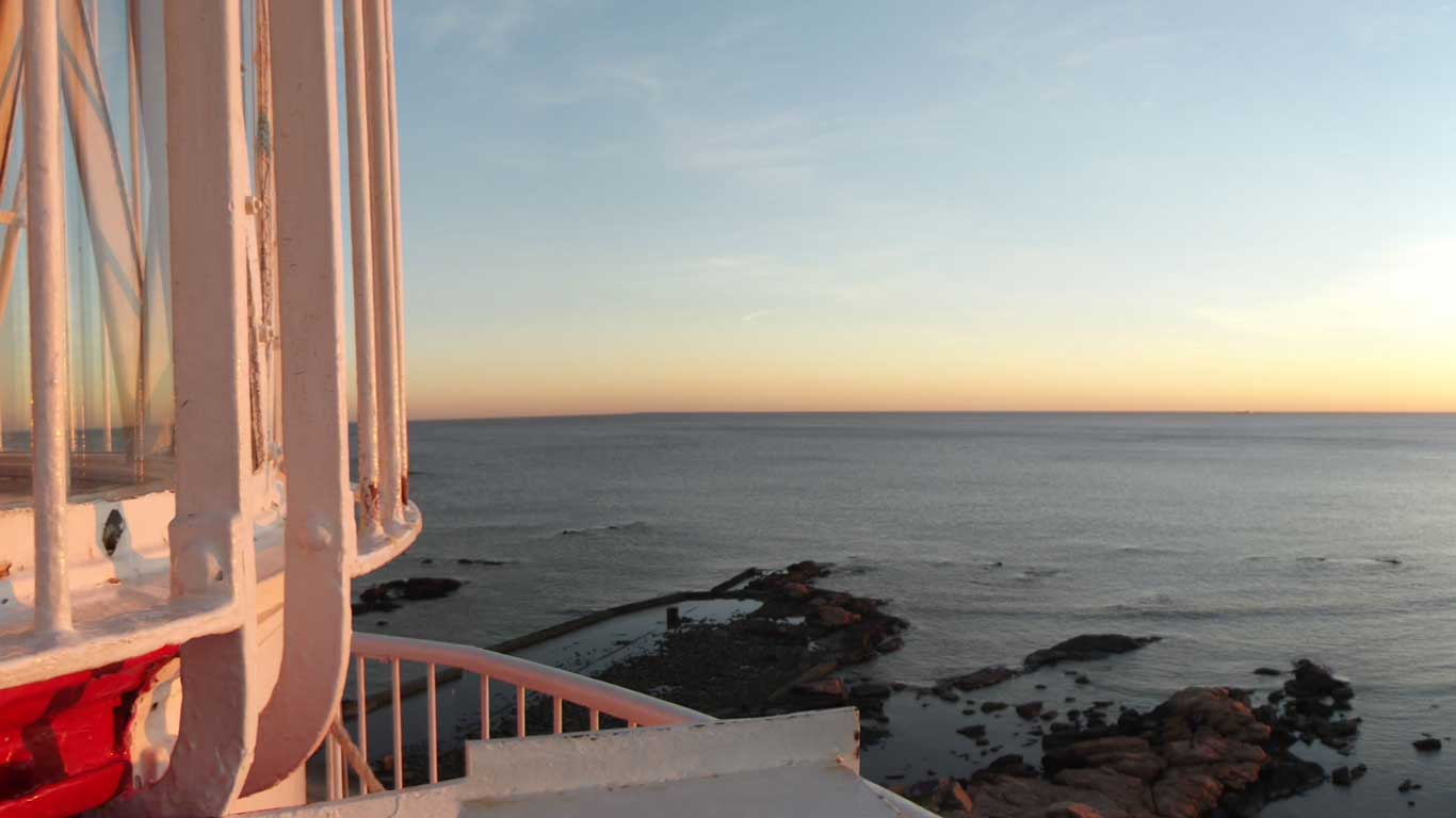 View from Punta Carretas Lighthouse in Montevideo, showing the lighthouse's structure on the left and a vast expanse of ocean at sunset, with the sky transitioning from light blue to warm orange hues.