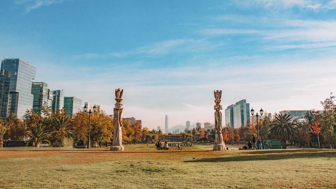 A panoramic view of a park in Las Condes, Santiago, with two large wooden sculptures and lush vegetation. Modern skyscrapers rise in the background, mixing nature with urban architecture, transforming the neighborhood into a great choice for where to stay in Santiago.