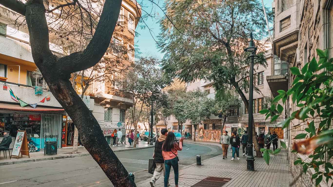 A lively street scene in the Lastarria neighborhood of Santiago, Chile, featuring people walking, chatting, and enjoying the vibrant atmosphere. The street is lined with trees, colorful buildings, and a variety of shops, adding to the charm of the area which is the best place to stay in Santiago, Chile.