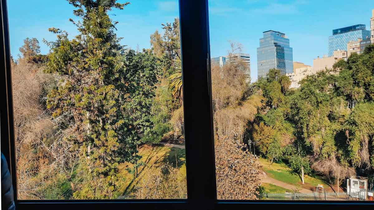 View from a window at the Foresta Hotel in Santiago, Chile, overlooking a park with lush green trees. Modern high-rise buildings are visible in the background under a clear blue sky. The scene captures the contrast between the urban skyline and the natural greenery.