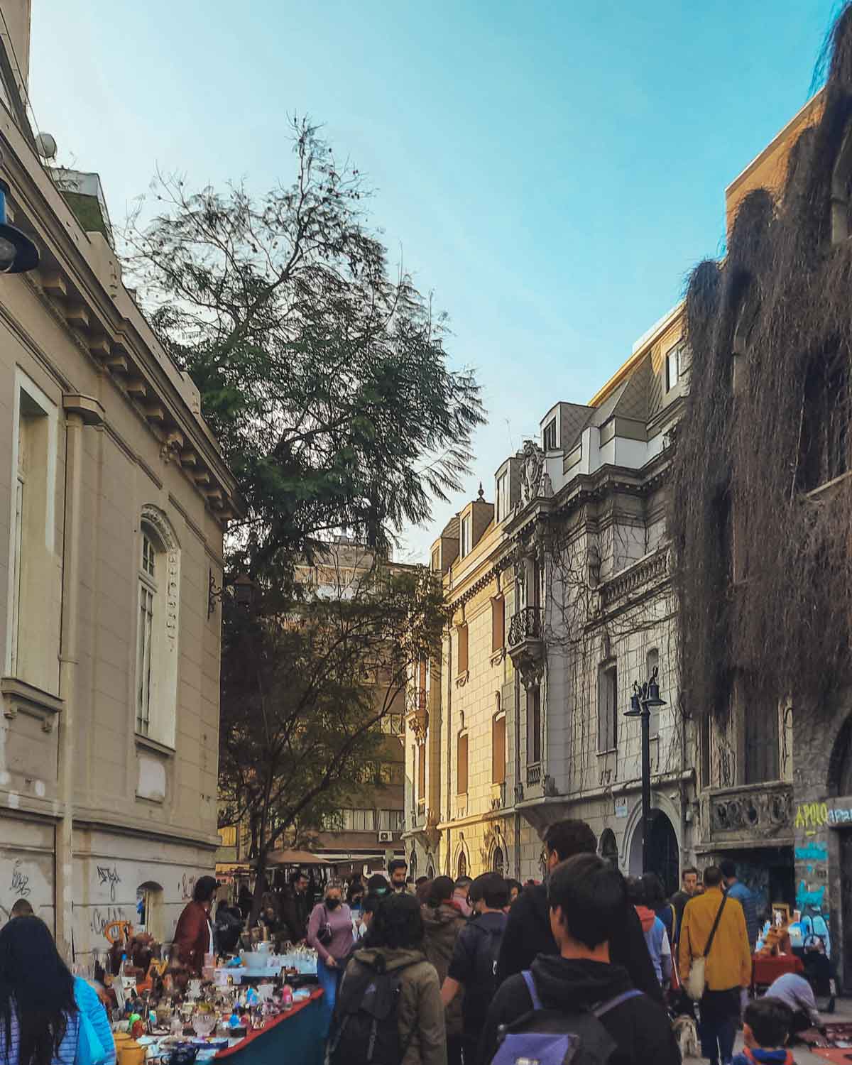 A bustling street market in the Lastarria neighborhood of Santiago, Chile. Vendors line the sides with tables displaying various goods while people walk through the narrow street. The scene is framed by historic buildings and a clear blue sky.