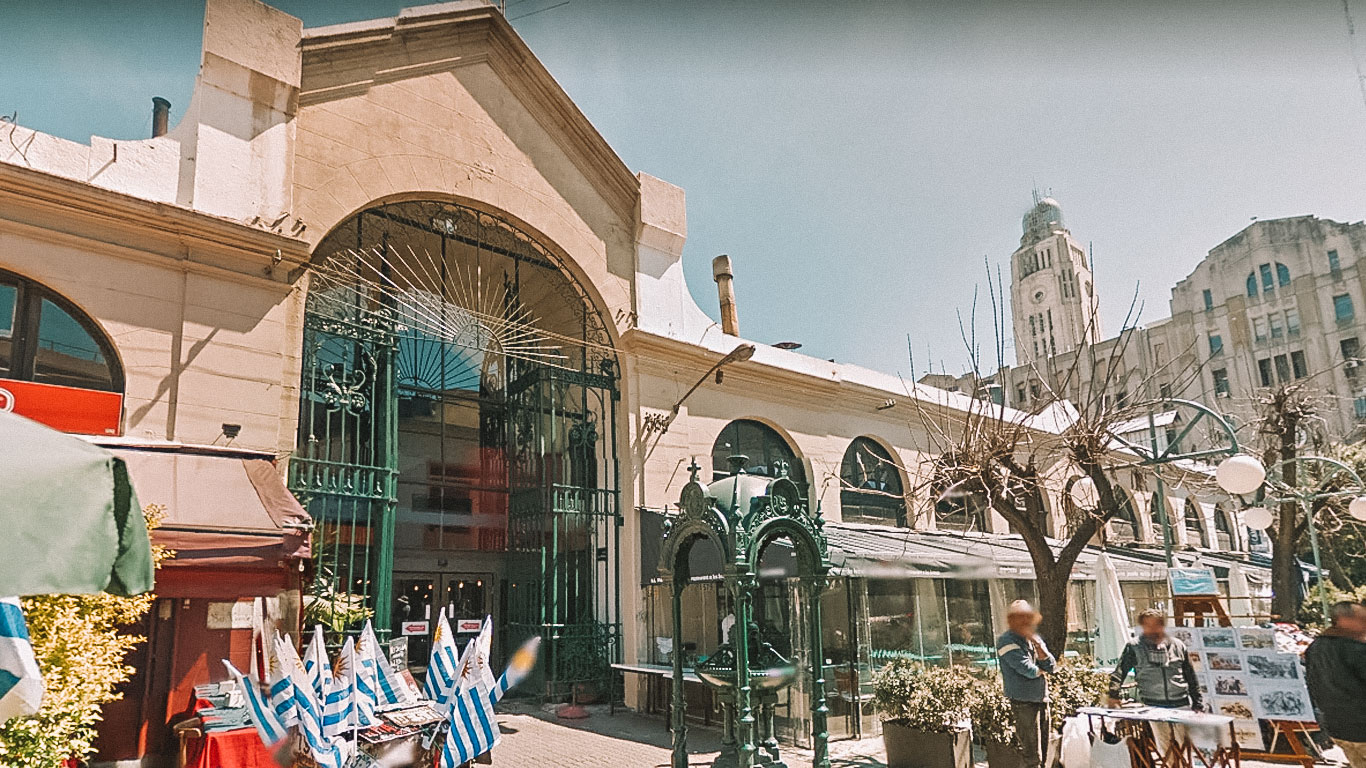 The entrance of Mercado del Puerto in Montevideo, featuring an arched doorway adorned with decorative metalwork. The market area is bustling with activity, showcasing stalls with Uruguayan flags and people engaging with vendors under a clear sky.