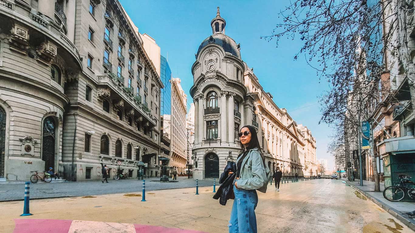 A woman stands on a street in downtown Santiago, Chile, with the historic Bolsa de Comercio building in the background. The scene captures a mix of classic architecture and a lively urban atmosphere under a clear blue sky.