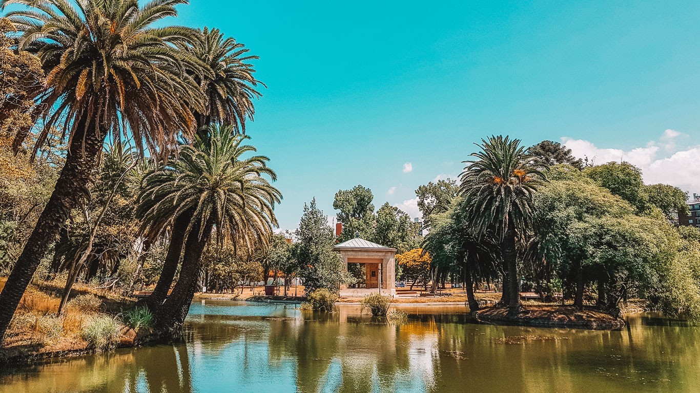 A picturesque scene at Parque Rodó in Montevideo, featuring tall palm trees and lush greenery surrounding a calm, reflective pond. In the background, a small, classical-style pavilion adds a charming architectural element to the vibrant landscape. The bright blue sky enhances the tranquil and inviting atmosphere of the park.