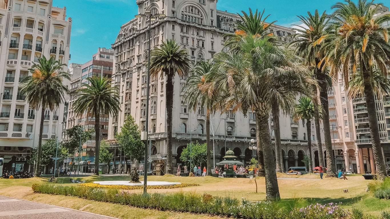 A vibrant view of Plaza Independencia in Montevideo, Uruguay, featuring lush green lawns, tall palm trees, and people enjoying the sunny day. The historic and ornate buildings in the background add a touch of architectural grandeur to the scene.