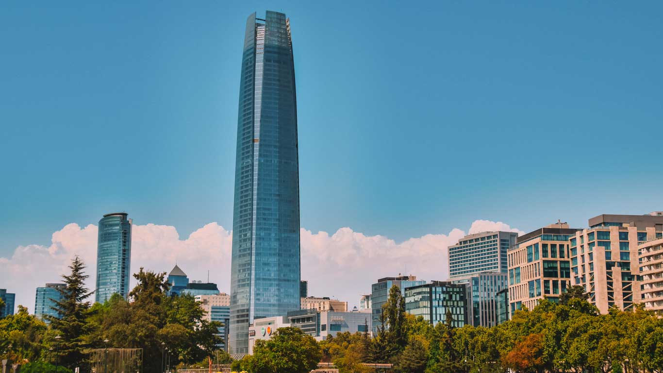 A clear view of the Gran Torre Santiago skyscraper in the Providencia neighborhood of Santiago, Chile, with modern buildings and lush greenery in the foreground. The bright blue sky and fluffy white clouds create a picturesque urban landscape.