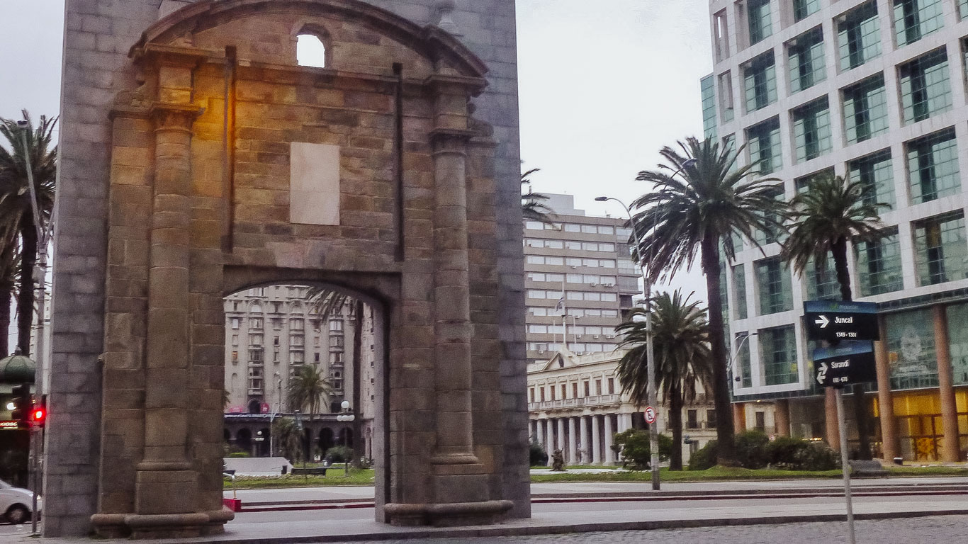 View of Puerta de la Ciudadela in Montevideo, Uruguay, with the historic stone arch framed by modern office buildings and palm trees. The scene includes street signs pointing to Juncal and Sarandí. A stroll around the square is one of the best things to do in Montevideo.