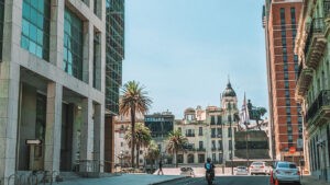 The image shows a street scene in Montevideo, featuring a mix of modern and historic architecture. On the left, there's a modern building with large glass windows, while in the center and right, historic buildings with ornate facades and palm trees are visible. In the background, there is a statue of a person on horseback, flanked by the Uruguayan flag.