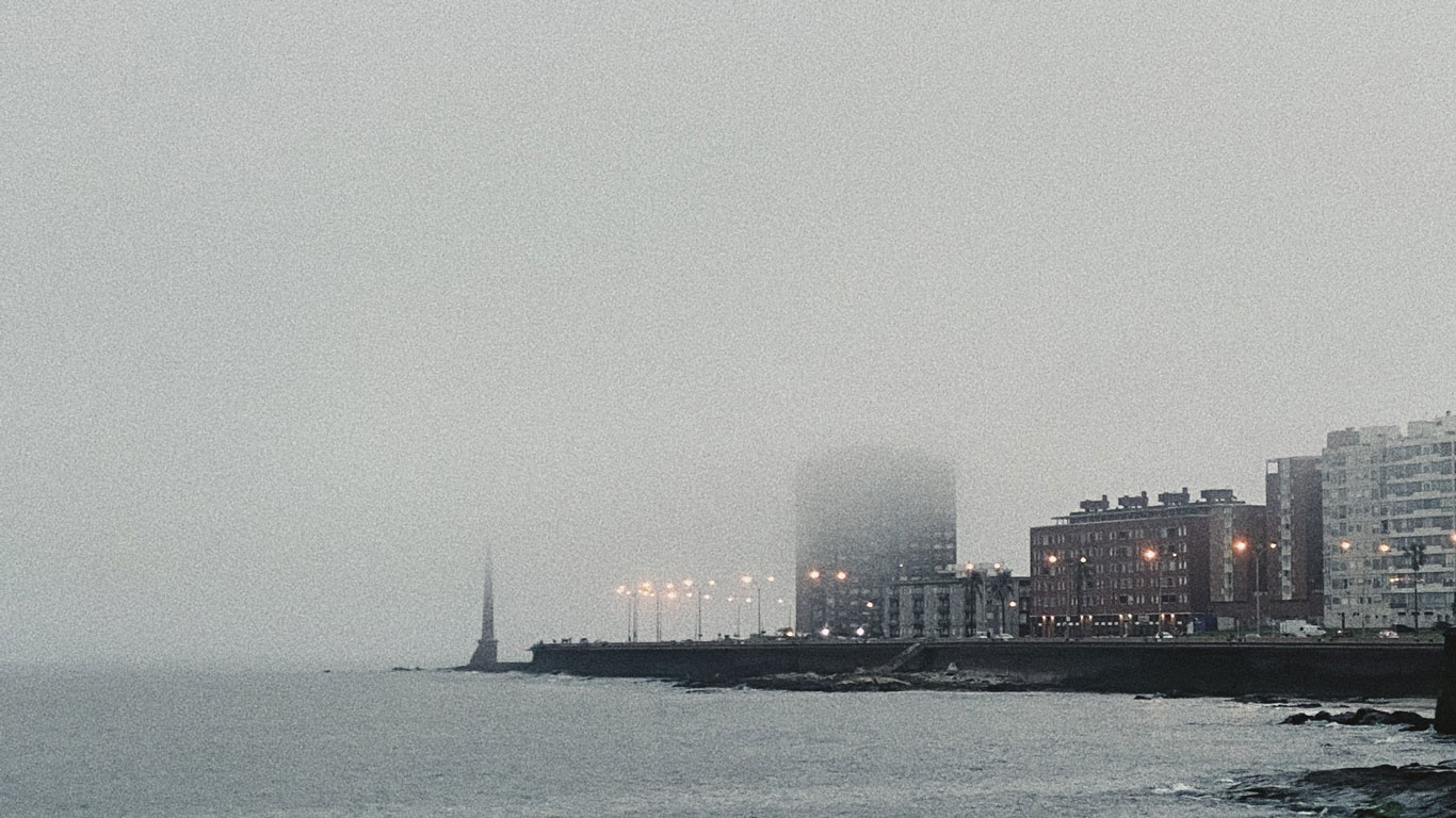 A foggy view of the Rambla of Montevideo, with buildings and streetlights partially obscured by the mist. The shoreline and water are visible in the foreground, creating a moody and atmospheric scene.