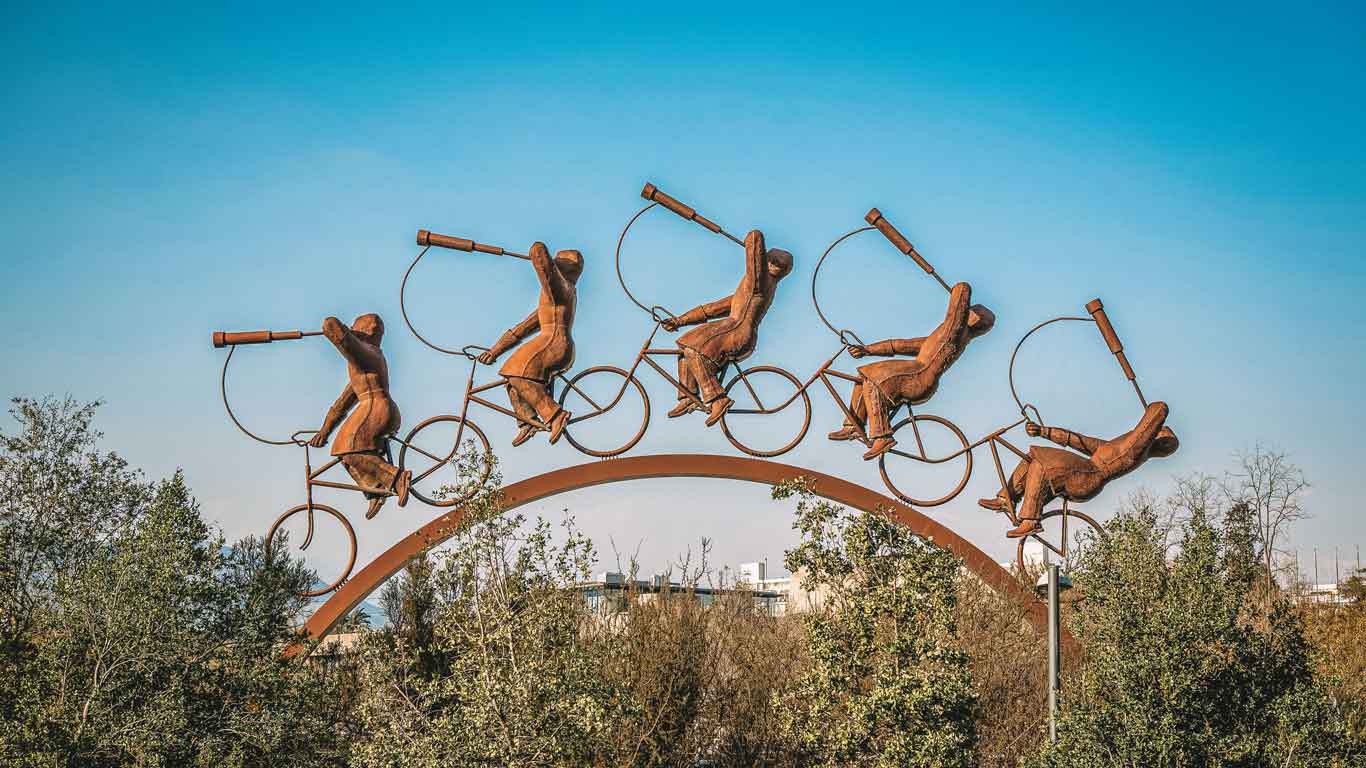 A whimsical sculpture in Vitacura, Santiago, featuring four figures riding bicycles on an arch, each holding a telescope. The artwork is set against a clear blue sky, with treetops in the foreground and urban buildings faintly visible in the background.