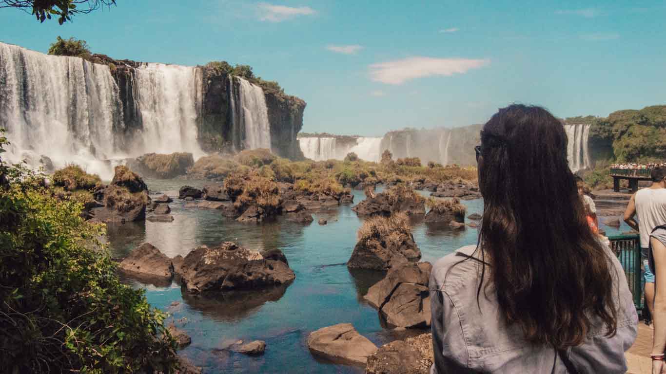 A woman with long hair gazing at the majestic Iguazu Falls in Foz do Iguaçu. The powerful waterfalls cascade over rocky cliffs into the river below, surrounded by lush greenery and clear blue skies.