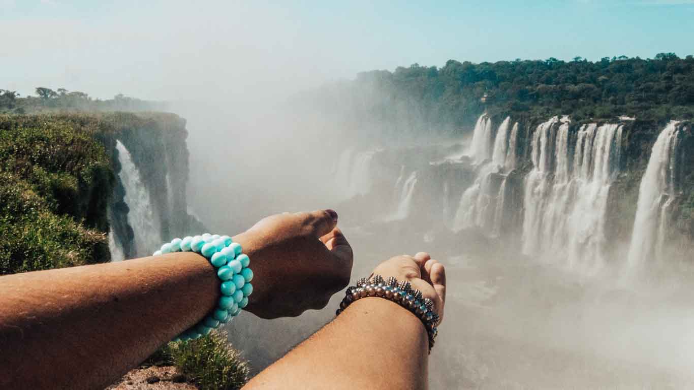 Close-up of two hands wearing bracelets, reaching out towards the misty expanse of Iguazu Falls in Puerto Iguazú. The powerful waterfalls cascade down the cliffs amidst lush greenery, creating a dramatic and awe-inspiring scene enveloped in mist.