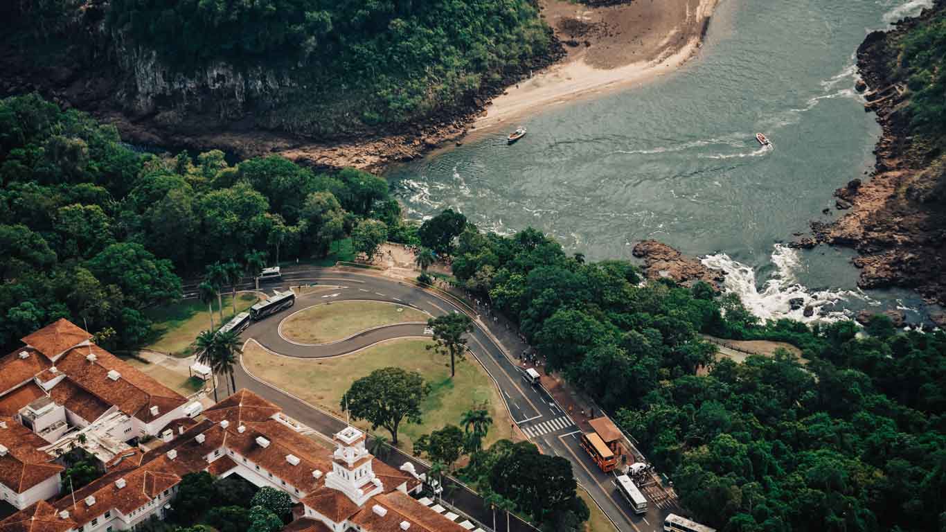 Aerial view of the Hotel das Cataratas located next to the winding Iguaçu River, an excellent option for where to stay when visiting Iguazu Falls. The hotel's red roofs and white buildings are surrounded by a lush green forest, with a curving road and parked buses visible nearby. 