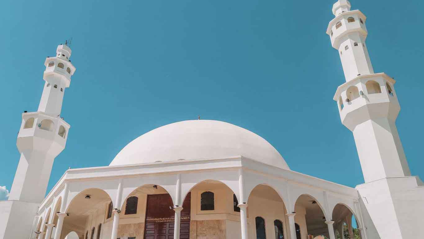 A bright, sunny view of a white mosque in Foz do Iguaçu, featuring two tall minarets and a large central dome. The mosque's elegant arches and clean lines stand out against the clear blue sky.