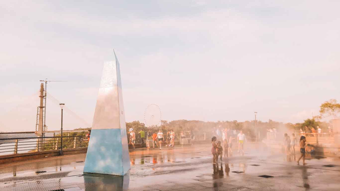 People enjoying a sunny day at the Hito de las Tres Fronteras in Puerto Iguazú, with children playing in the mist of a water fountain. The landmark obelisk painted in the colors of the Argentine flag stands prominently, with a ferris wheel and bridge visible in the background.