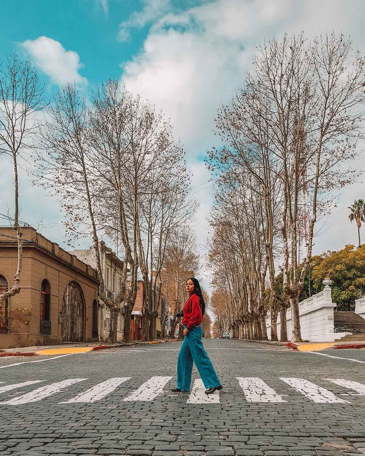 A woman with long hair, wearing blue jeans, boots, and a red hoodie, crossing the pedestrian crosswalk in the old town of Colonia del Sacramento. With colonial houses in the background and a street lined with leafless trees due to winter, fading into the horizon.