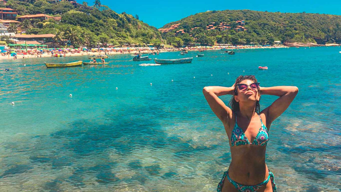 A woman in a bikini enjoys the sun and crystal-clear water at João Fernandes Beach in Búzios. In the background, the beach is filled with sunbathers under colorful umbrellas and boats floating near the shore, surrounded by green hills.