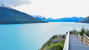 The image shows a breathtaking view of a turquoise lake surrounded by lush green mountains with snow-capped peaks in the distance. A wooden walkway with railings descends towards the lake, inviting exploration of the natural landscape. The sky is mostly cloudy, creating a serene and peaceful atmosphere.