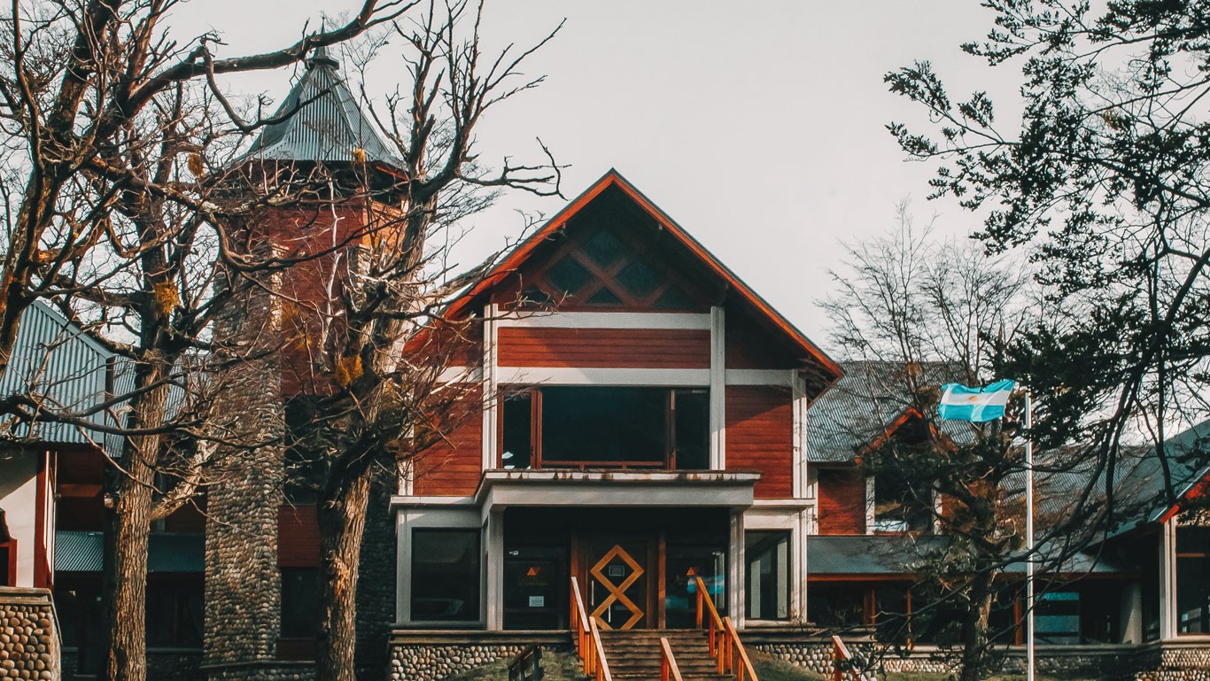 The image depicts a charming and rustic building with red wooden siding and a stone foundation, set against a backdrop of leafless trees. The building features a large central entrance with a steeply pointed gable roof and a tower-like structure beside it—an excellent place where to stay in El Calafate.