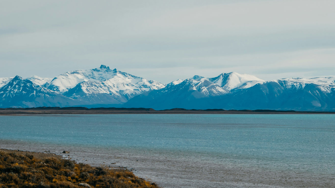 The image shows a vast, tranquil body of water in the foreground with a distant shoreline leading up to majestic snow-capped mountains. The mountains have rugged peaks and are partially shrouded in mist or clouds, creating a serene and awe-inspiring scene in El Calafate, Argentina.