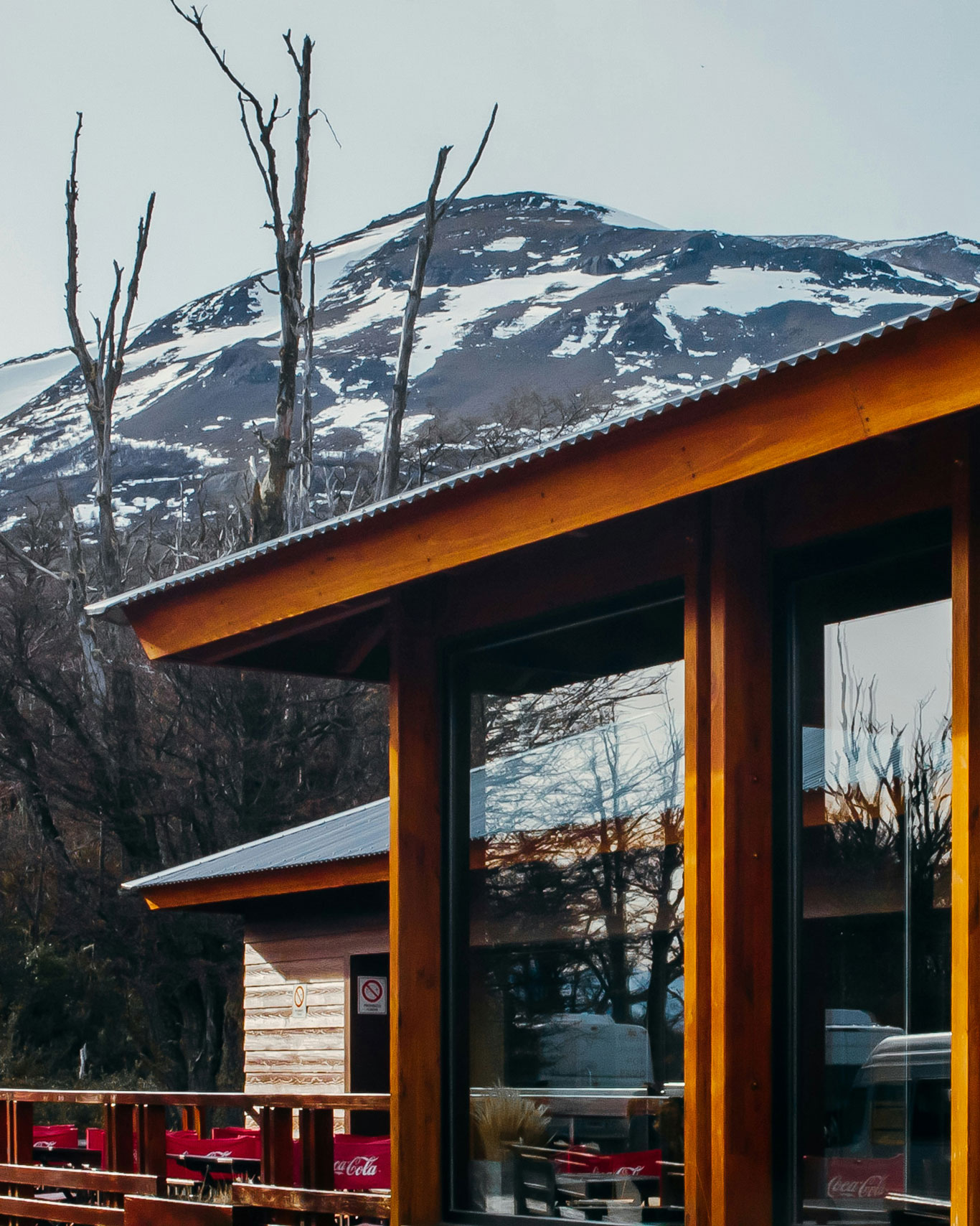 The image depicts a rustic wooden building with large windows reflecting the surrounding landscape. In the background, a snow-covered mountain rises under a pale sky. The setting appears to be in a serene, mountainous region, the best place to stay in El Calafate.