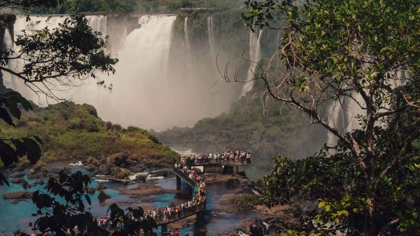 A breathtaking view of Iguazu Falls with a crowd of tourists walking along a curved pathway, surrounded by lush greenery and mist from the cascading waterfalls in the background.