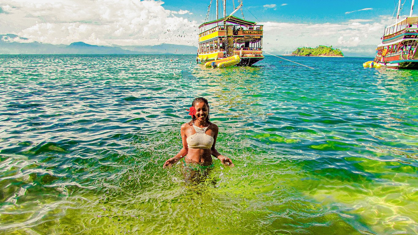 A woman with a red flower in her hair splashes water upwards with her hands at a beach in Paraty, where the green waters meet the horizon. In the background, a schooner blends into the blue sky on the horizon.