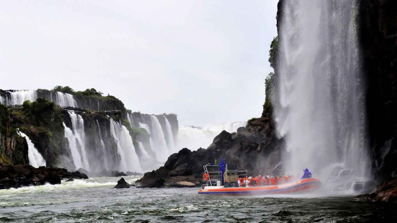 A boat filled with tourists in orange life vests approaching the powerful cascade of Iguazu Falls, with water misting around them and the dramatic waterfalls towering in the background.