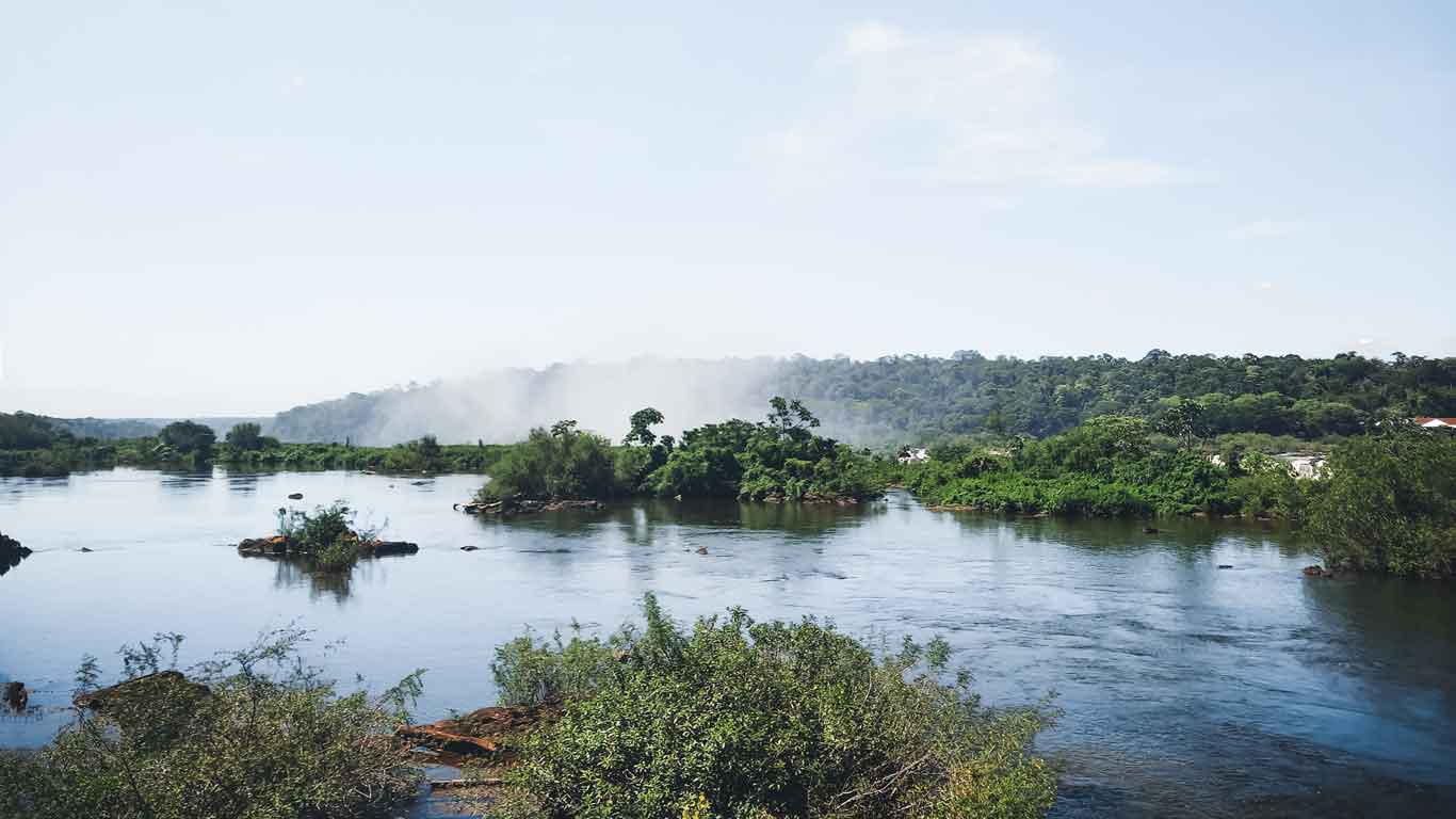 A serene river landscape leading up to Iguazu Falls, with mist rising in the distance and lush greenery lining the water's edge under a clear blue sky.