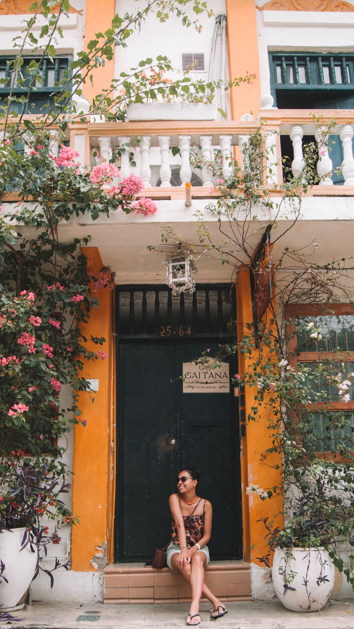 The image shows a woman seated on the steps of a vibrant, colonial-style doorway in Cartagena, framed by lush bougainvillea and other green plants. The orange and white facade complements the deep green door, which is labeled "Casa Gaitana." This scene captures a relaxed, picturesque moment, evoking the historic and colorful charm of Cartagena's streets.